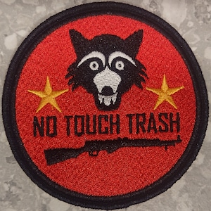 May include: A red and black embroidered patch featuring a raccoon face, two yellow stars, and the text "NO TOUCH TRASH" with a black rifle underneath.