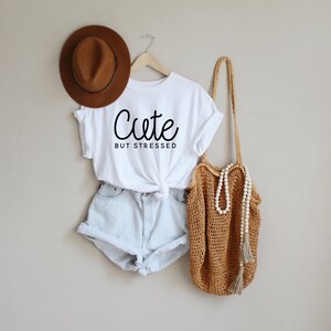 May include: White t-shirt with the text "Cute but stressed" in black cursive writing. The shirt is paired with light blue denim shorts and a brown crocheted tote bag with a white beaded necklace.