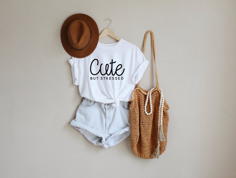 May include: White t-shirt with the text "Cute but stressed" in black cursive writing. The shirt is paired with light blue denim shorts and a brown crocheted tote bag with a white beaded necklace.