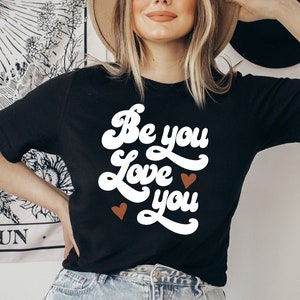 May include: Black t-shirt with white text that reads "Be you Love you" in a retro font. Two small brown hearts are on either side of the text.