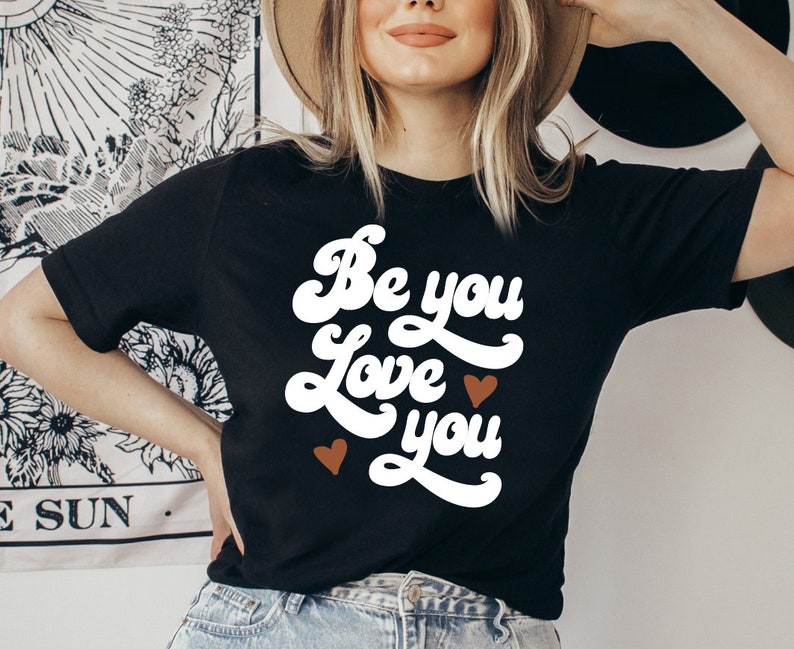 May include: Black t-shirt with white text that reads "Be you Love you" in a retro font. Two small brown hearts are on either side of the text.