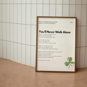 Könnte beinhalten: Ein gerahmter Druck der Liedtexte zu dem Song "You'll Never Walk Alone" von Gerry and the Pacemakers. Die Texte sind in schwarzer Schrift auf weißem Hintergrund gedruckt. Der Rahmen ist aus braunem Holz und der Druck enthält ein grünes Kleeblatt.