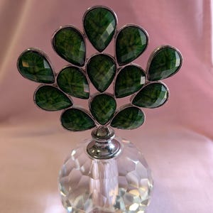 May include: A clear glass perfume bottle with a faceted design and a silver top. The top is decorated with a cluster of green, teardrop-shaped stones.