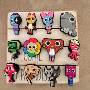 May include: A set of 12 colorful cartoon character shoe charms. The charms are made of rubber and feature various characters with different expressions and designs. The charms are displayed on a white wooden surface with the words "Welcome to Paradise" written on it.