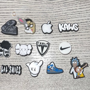 May include: A collection of 12 colorful shoe charms featuring various logos and characters, including a black and white rose, a black and white skull, a white apple logo, a white Tesla logo, a white Nike swoosh logo, a white Kaws logo, a white hand with an 'XX' on it, a white cartoon character with a blue hat, a white cartoon character with a blue hat and a red shirt, a white cartoon character with a blue hat and a red shirt, a white cartoon character with a blue hat and a red shirt, and a white cartoon character with a blue hat and a red shirt.