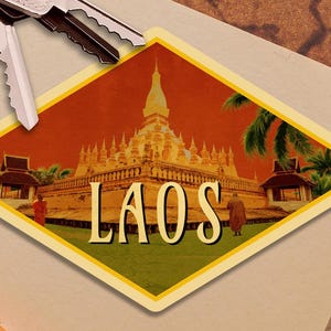 May include: A diamond-shaped sticker featuring a golden temple with the word "LAOS" in large, cream-colored letters. The background is a warm orange, with palm trees and figures. Silver keys rest above the sticker.