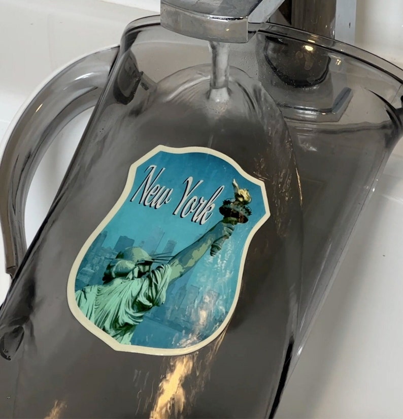 May include: A gray glass pitcher with a sticker of the Statue of Liberty and the words "New York" on it. The pitcher is being filled with water from a faucet.