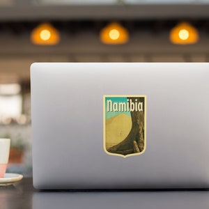 K&ouml;nnte beinhalten: Ein Aufkleber im Vintage-Stil mit einem Farbschema in Gelb, Orange und Braun. Der Aufkleber zeigt eine gro&szlig;e Sandd&uuml;ne mit einem Baum im Hintergrund. Der Text "Namibia" ist in fetter, schwarzer Schrift gedruckt.