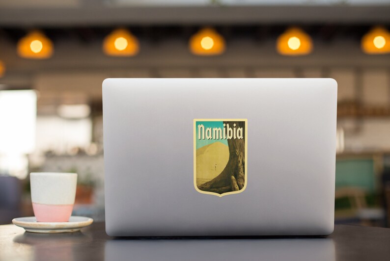 K&ouml;nnte beinhalten: Ein Aufkleber im Vintage-Stil mit einem Farbschema in Gelb, Orange und Braun. Der Aufkleber zeigt eine gro&szlig;e Sandd&uuml;ne mit einem Baum im Hintergrund. Der Text "Namibia" ist in fetter, schwarzer Schrift gedruckt.