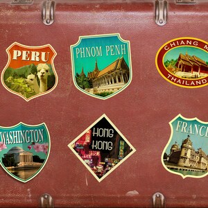 May include: A vintage suitcase covered in travel stickers. The stickers feature destinations like Peru, Phnom Penh, Chiang Mai, Thailand, Washington, Hong Kong, and France.