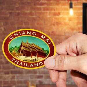 May include: A round, red and gold sticker with the text "CHIANG MAI THAILAND" in a yellow circle. The sticker features a vintage illustration of a temple with a blue sky and green trees.