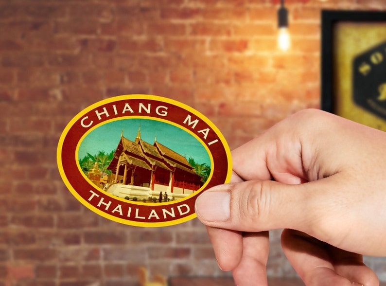 May include: A round, red and gold sticker with the text "CHIANG MAI THAILAND" in a yellow circle. The sticker features a vintage illustration of a temple with a blue sky and green trees.