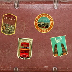 May include: A brown vintage suitcase with four travel stickers. The stickers are for Porto, Portugal; San Francisco, California; London, England; and Bali, Indonesia.