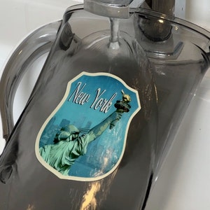 May include: A gray glass pitcher with a handle, filled with water. A sticker with a blue background and the text "New York" is on the side of the pitcher. The sticker features a depiction of the Statue of Liberty.