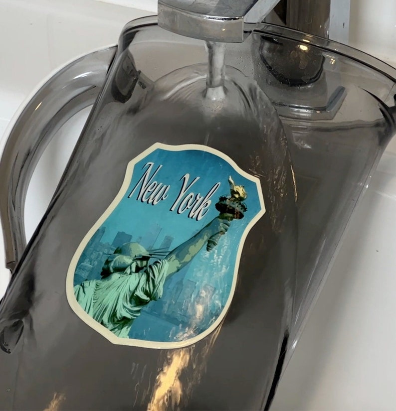 May include: A gray glass pitcher with a handle, filled with water. A sticker with a blue background and the text "New York" is on the side of the pitcher. The sticker features a depiction of the Statue of Liberty.