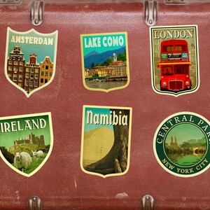K&ouml;nnte beinhalten: Ein Vintage-Koffer mit Reiseaufklebern aus Amsterdam, Comer See, London, Irland, Namibia und Central Park, New York City.