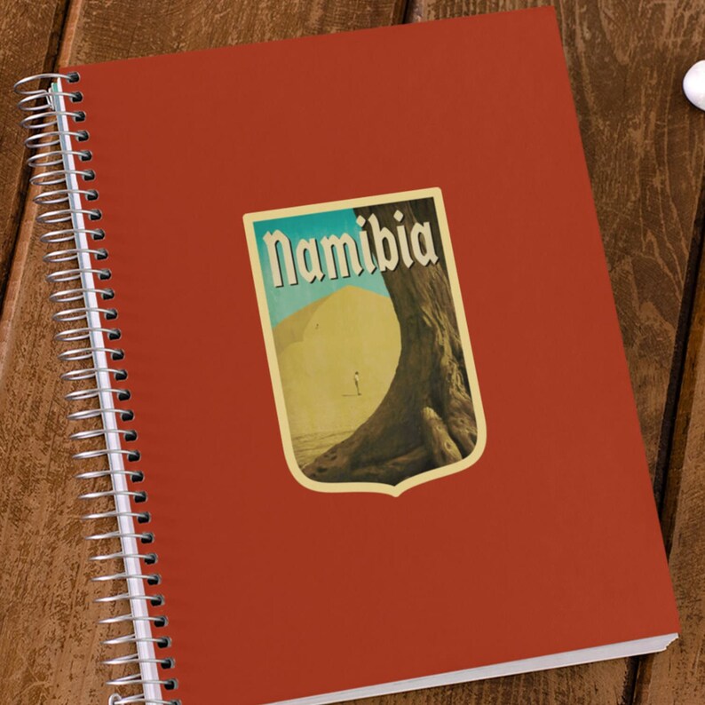 K&ouml;nnte beinhalten: Ein rotes Spiralnotizbuch mit einem Vintage-Reisesticker, der eine W&uuml;stenlandschaft und das Wort "Namibia" in einer stilisierten Schrift zeigt.