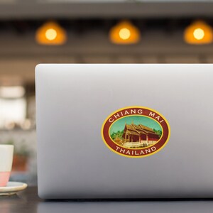 May include: A laptop computer with a sticker of a red building with a yellow roof and the text "CHIANG MAI THAILAND" in a circle.