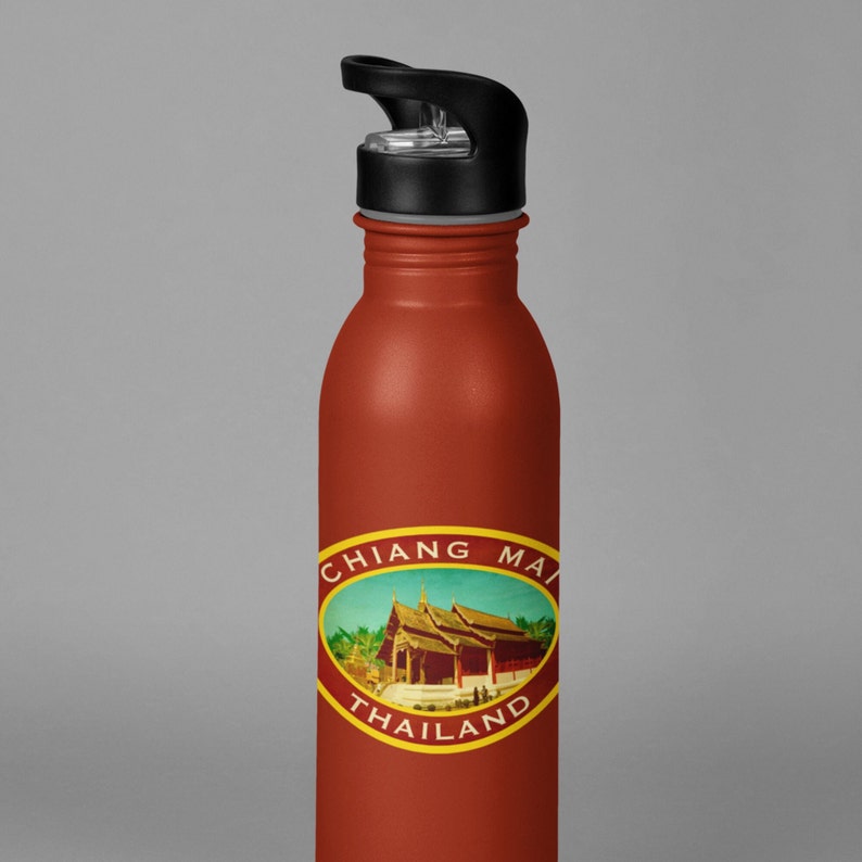 May include: A red stainless steel water bottle with a black lid and a white oval sticker that says "Chiang Mai Thailand" with a picture of a building in the center.