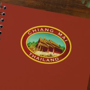 May include: A vintage-style travel sticker with a yellow border and the text "CHIANG MAI THAILAND". The sticker features a colorful image of a traditional Thai temple with a blue sky and green trees.