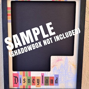 May include: Black shadowbox frame with a colorful Disneyland design. The frame has a black border and a black interior. The Disneyland text is in colorful block letters. The image includes the text "SAMPLE (SHADOWBOX NOT INCLUDED)".