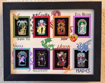 May include: A framed collection of enamel pins featuring Disney villains. The pins are displayed in individual frames with character names like Cruella, Evil Queen, and Hades. The frame is black, and the background is a light fabric.