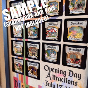 May include: A framed display of Disneyland Resort pins, featuring various attraction designs. The display includes the text "Opening Day Attractions July 17, 1955" and the Disneyland logo. The frame is black with a colorful striped background.