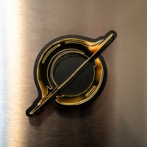 May include: A gold-colored metal logo with a circular design and a diagonal line through it. The logo is on a black background.