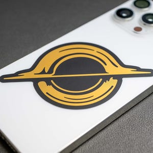 May include: A gold and black sticker with a stylized image of a black hole. The sticker is on a white iPhone.