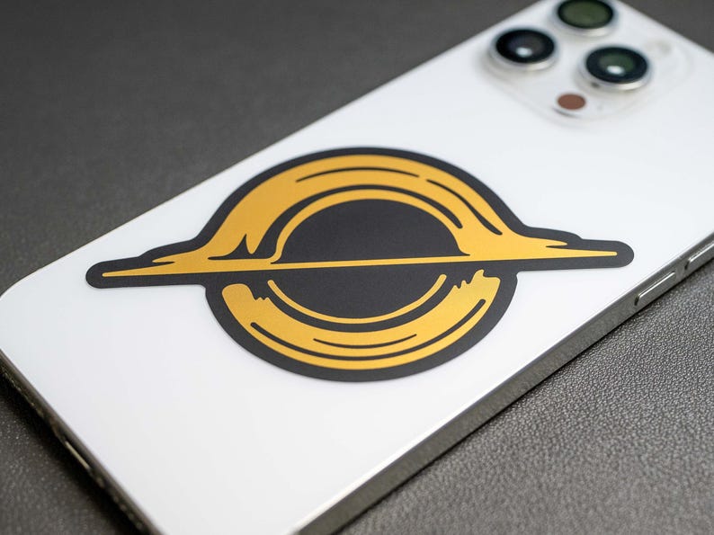 May include: A gold and black sticker with a stylized image of a black hole. The sticker is on a white iPhone.