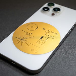May include: A white iPhone with a gold sticker of the Voyager Golden Record on the back. The sticker features a stylized image of the record, a diagram of the record's contents, and a diagram of the solar system.