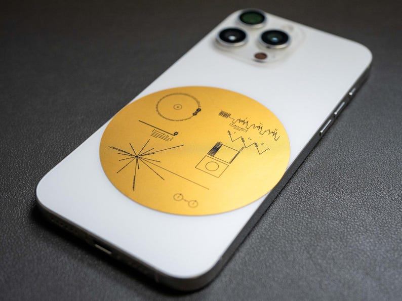 May include: A white iPhone with a gold sticker of the Voyager Golden Record on the back. The sticker features a stylized image of the record, a diagram of the record's contents, and a diagram of the solar system.