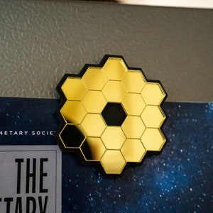 May include: A gold hexagonal mirror with a black centre, resembling the James Webb Space Telescope. The mirror is on a grey surface with a blue background.