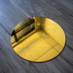 May include: A gold-colored, circular, metal disc with a design etched into its surface. The design includes a series of lines radiating outward from a central point, along with other geometric shapes and symbols. The disc is likely a replica of the Voyager Golden Record, which was sent into space on the Voyager spacecraft.