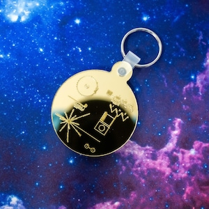 May include: A gold-colored keychain with a round, mirrored disc featuring a stylized image of a spacecraft, a record, and a diagram of a human figure. The disc is surrounded by a silver key ring.