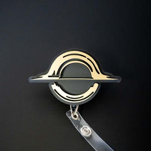 May include: A black and gold badge reel featuring a stylized black hole design. The badge has a gold outline and accents, with a clear plastic strap and a metal clip for attaching to clothing or accessories. The background is black.
