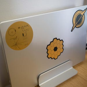 May include: A silver laptop computer with a stand. The laptop has three stickers on it: a yellow circle with a black design, a black and yellow honeycomb design, and a black and white design of a ring.