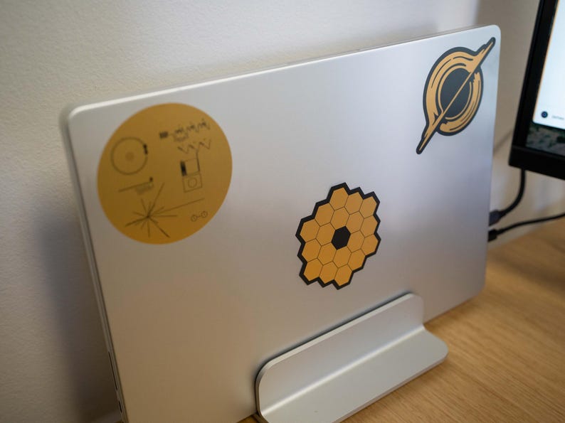 May include: A silver laptop computer with a stand. The laptop has three stickers on it: a yellow circle with a black design, a black and yellow honeycomb design, and a black and white design of a ring.