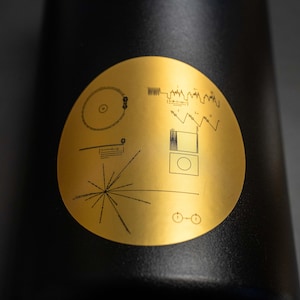 May include: A black water bottle with a gold sticker featuring a stylized image of the Voyager Golden Record, a diagram of the solar system, and a series of lines representing the hydrogen atom.