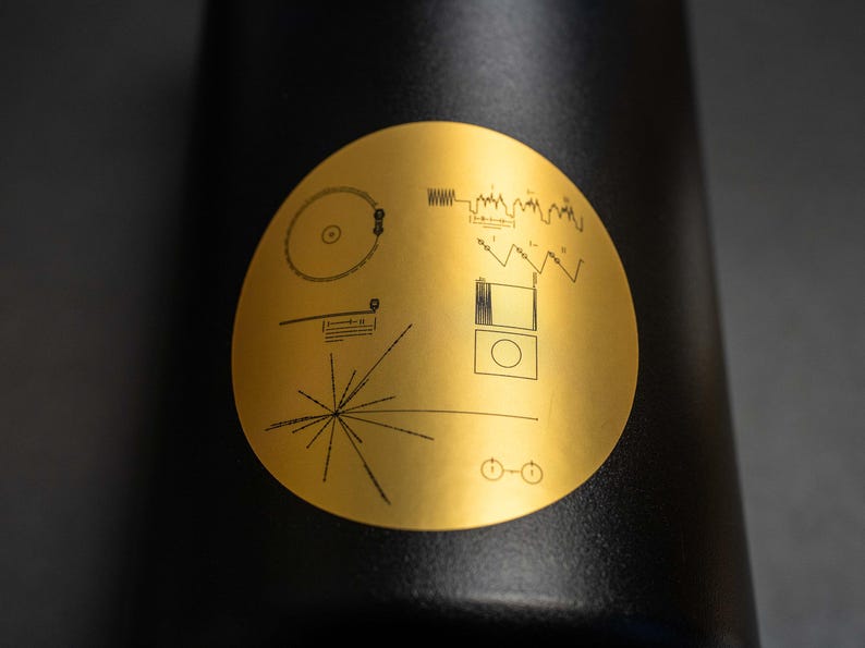 May include: A black water bottle with a gold sticker featuring a stylized image of the Voyager Golden Record, a diagram of the solar system, and a series of lines representing the hydrogen atom.
