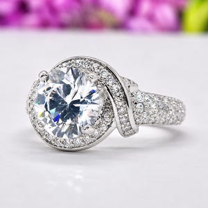 Puede incluir: Un anillo plateado con una piedra preciosa grande, redonda y transparente en el centro, rodeada de piedras m&aacute;s peque&ntilde;as y brillantes. La banda tambi&eacute;n est&aacute; incrustada con piedras m&aacute;s peque&ntilde;as, creando un efecto deslumbrante. El anillo est&aacute; sobre un fondo suave y borroso.