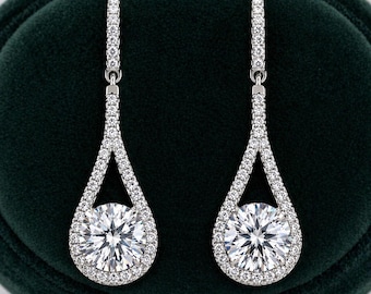 May include: A pair of elegant drop earrings. Each earring features a large, round, clear gemstone surrounded by smaller, sparkling stones. The earrings are set in a silver-colored metal and have a teardrop design, creating a sophisticated and eye-catching look.