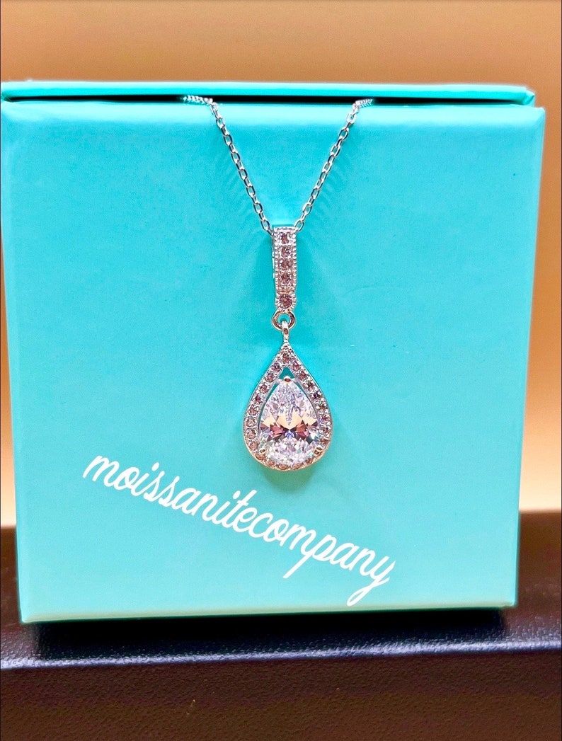 May include: A silver necklace with a pear-shaped pendant set with a large clear stone surrounded by smaller clear stones. The necklace is in a light blue box with the text "moissanitecompany" printed on the bottom.