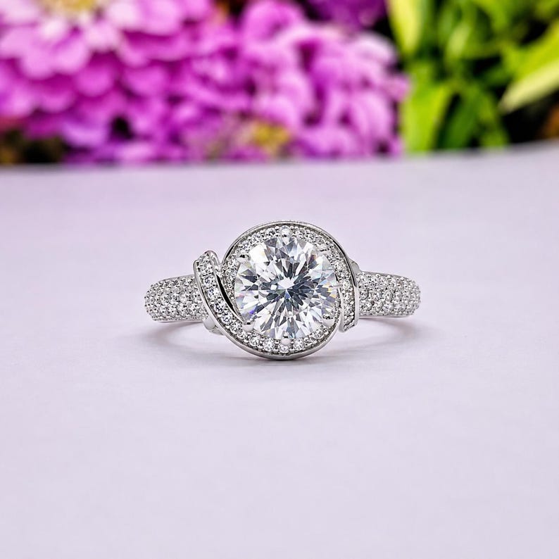 Puede incluir: Un anillo plateado con una piedra preciosa grande, redonda y transparente en el centro, rodeada de piedras m&aacute;s peque&ntilde;as. La banda tambi&eacute;n est&aacute; incrustada con peque&ntilde;as piedras. El anillo se presenta sobre un fondo morado suave, sugiriendo un tema de joyer&iacute;a.