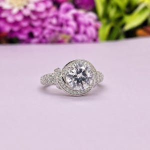 Puede incluir: Un anillo plateado con una piedra preciosa grande, redonda y transparente en el centro, rodeada de piedras m&aacute;s peque&ntilde;as. La banda y el halo circundante tambi&eacute;n est&aacute;n incrustados con piedras m&aacute;s peque&ntilde;as y brillantes. El anillo est&aacute; sobre un fondo morado suave.
