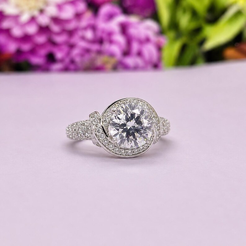 Puede incluir: Un anillo plateado con una piedra preciosa grande, redonda y transparente en el centro, rodeada de piedras m&aacute;s peque&ntilde;as. La banda y el halo circundante tambi&eacute;n est&aacute;n incrustados con piedras m&aacute;s peque&ntilde;as y brillantes. El anillo est&aacute; sobre un fondo morado suave.