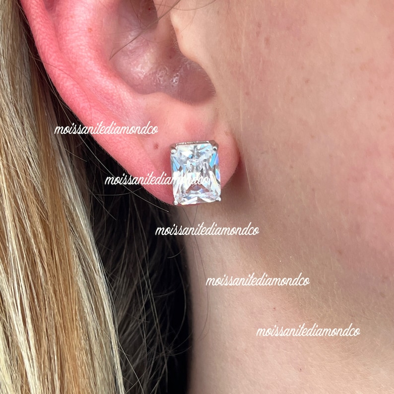 May include: A pair of square-shaped, clear, cubic zirconia earrings. The earrings are set in silver and are shown on a person's earlobe.