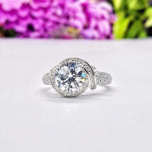 Puede incluir: Anillo plateado con una piedra preciosa grande, redonda y transparente como punto focal. La piedra preciosa est&aacute; rodeada por un halo de piedras m&aacute;s peque&ntilde;as. La banda presenta piedras peque&ntilde;as adicionales y un dise&ntilde;o de remolino decorativo.