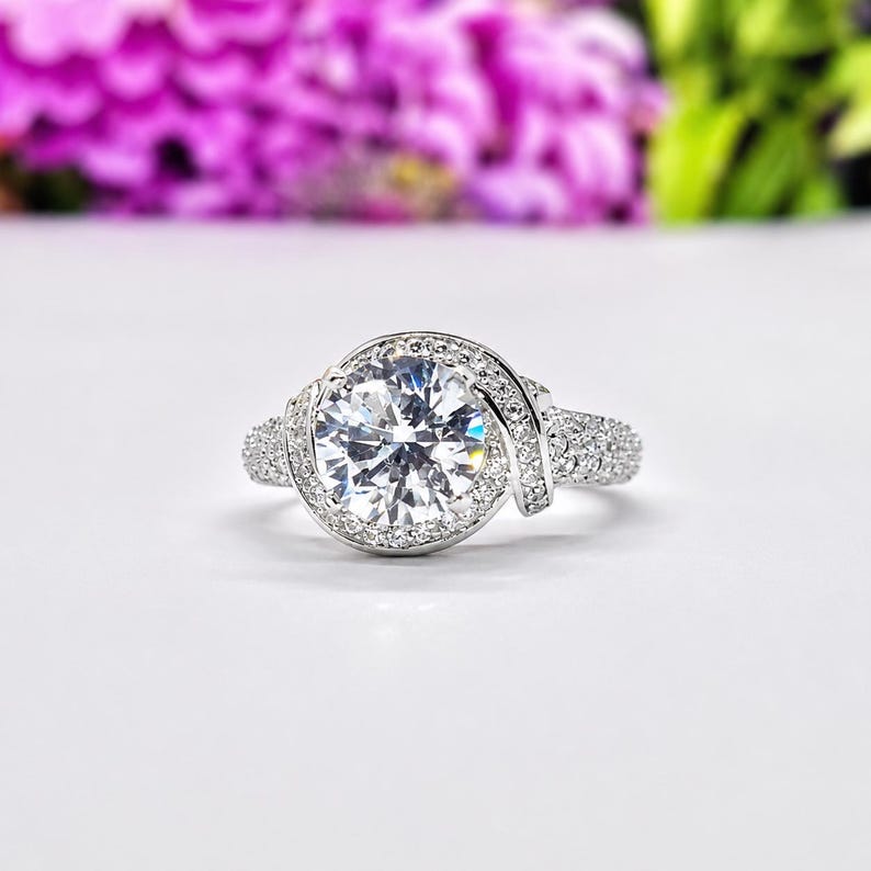 Puede incluir: Anillo plateado con una piedra preciosa grande, redonda y transparente como punto focal. La piedra preciosa est&aacute; rodeada por un halo de piedras m&aacute;s peque&ntilde;as. La banda presenta piedras peque&ntilde;as adicionales y un dise&ntilde;o de remolino decorativo.