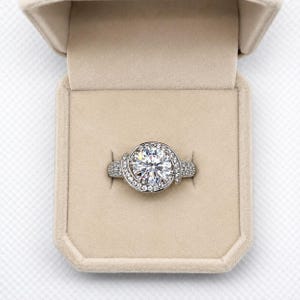 Puede incluir: Un anillo plateado con una piedra preciosa grande, redonda y transparente en el centro, rodeada de piedras m&aacute;s peque&ntilde;as y transparentes. El anillo est&aacute; en una caja beige.